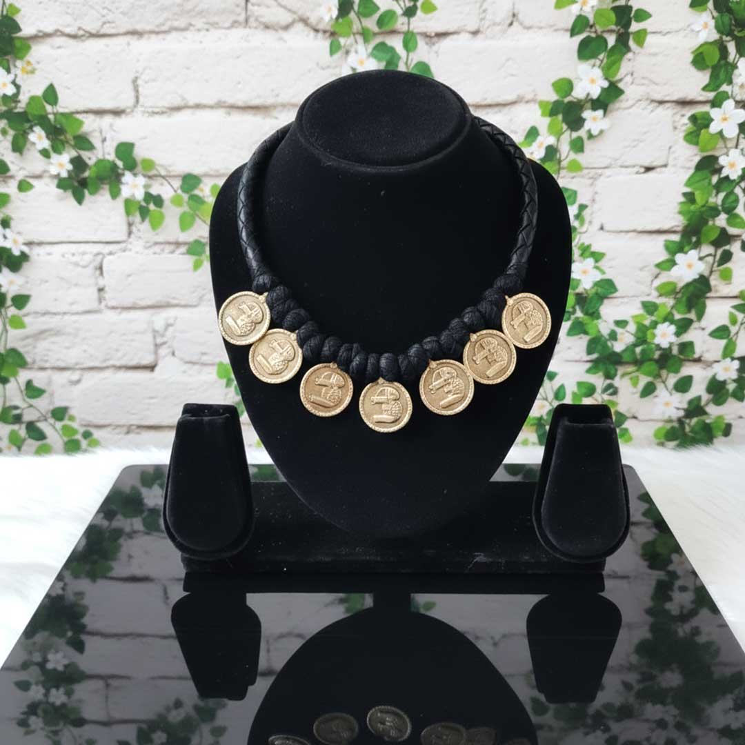 Black necklace with gold coins on a black stand against a white brick wall with greenery.
