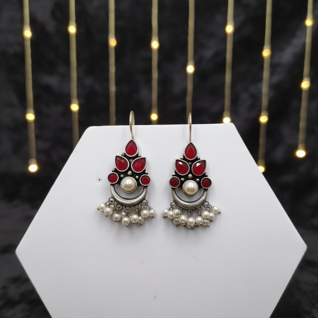 Pair of earrings with red stones and pearls on a white hexagonal stand with a dark background.
