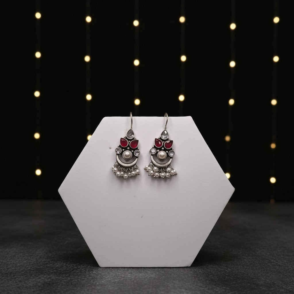 Pair of silver earrings with red stones on a white hexagonal stand against a dark background with string lights.
