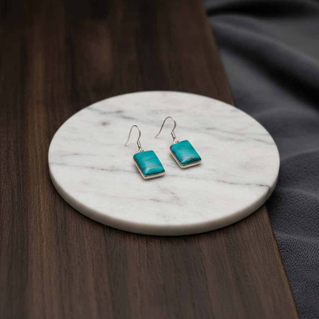 Turquoise square earrings on a marble coaster with a wooden surface.